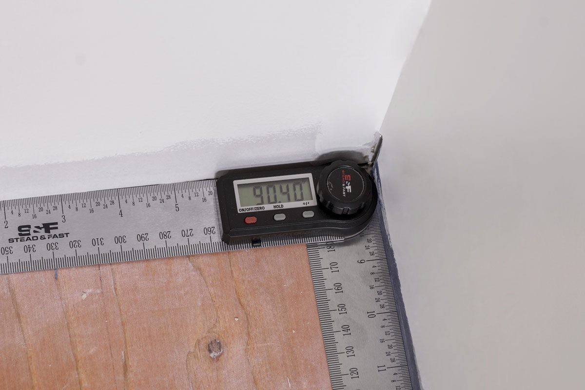 Overhead shot of a digital angle finder shows a reading of 94.40 degrees at the corner where a wall meets a wooden floor, with a metal ruler placed along both surfaces;
