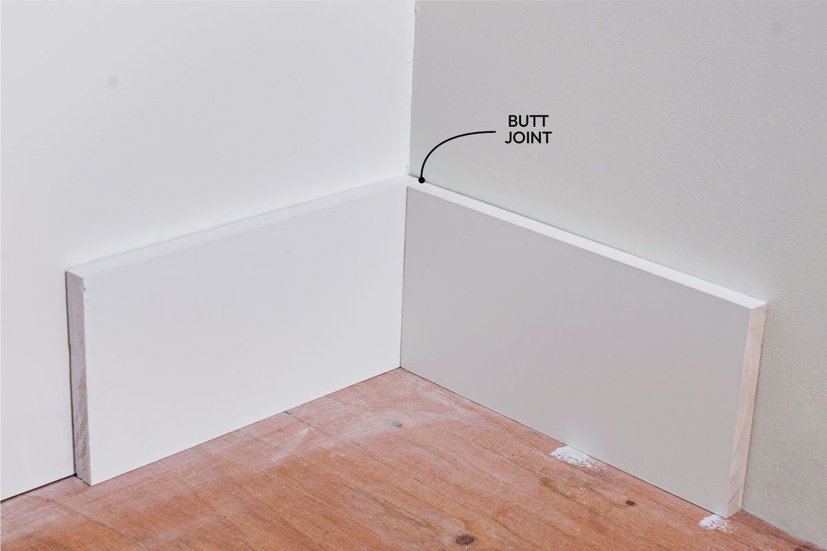 Close shot of two white baseboards are being installed at the corner of a room with light-colored walls and a wooden floor; One baseboard is placed along each wall, meeting at the corner;