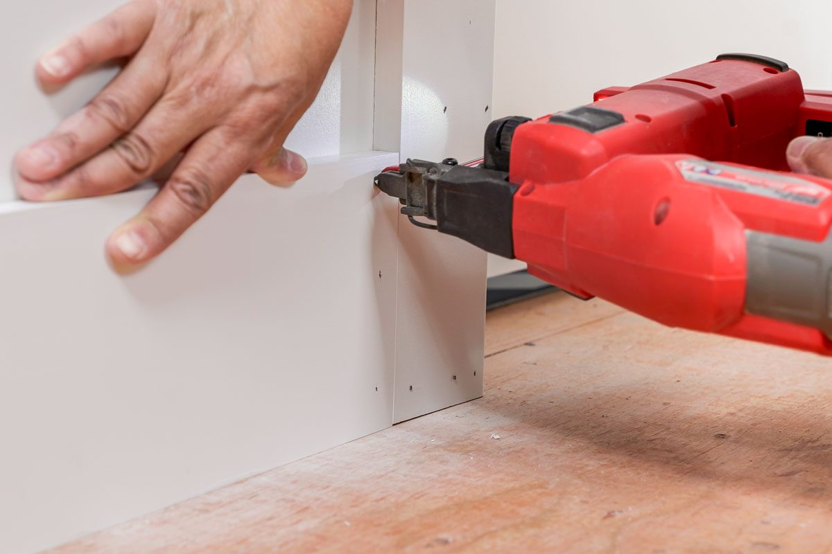 A person uses a red power nail gun to attach two white boards on a wooden floor; one hand steadies the boards