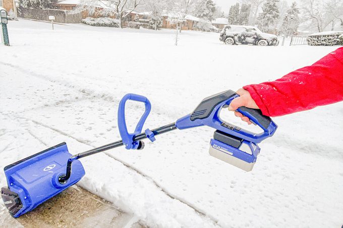 A person in a red jacket uses a blue cordless snow shovel to clear snow from a sidewalk on a snowy day; houses and a car are visible in the snowy background.