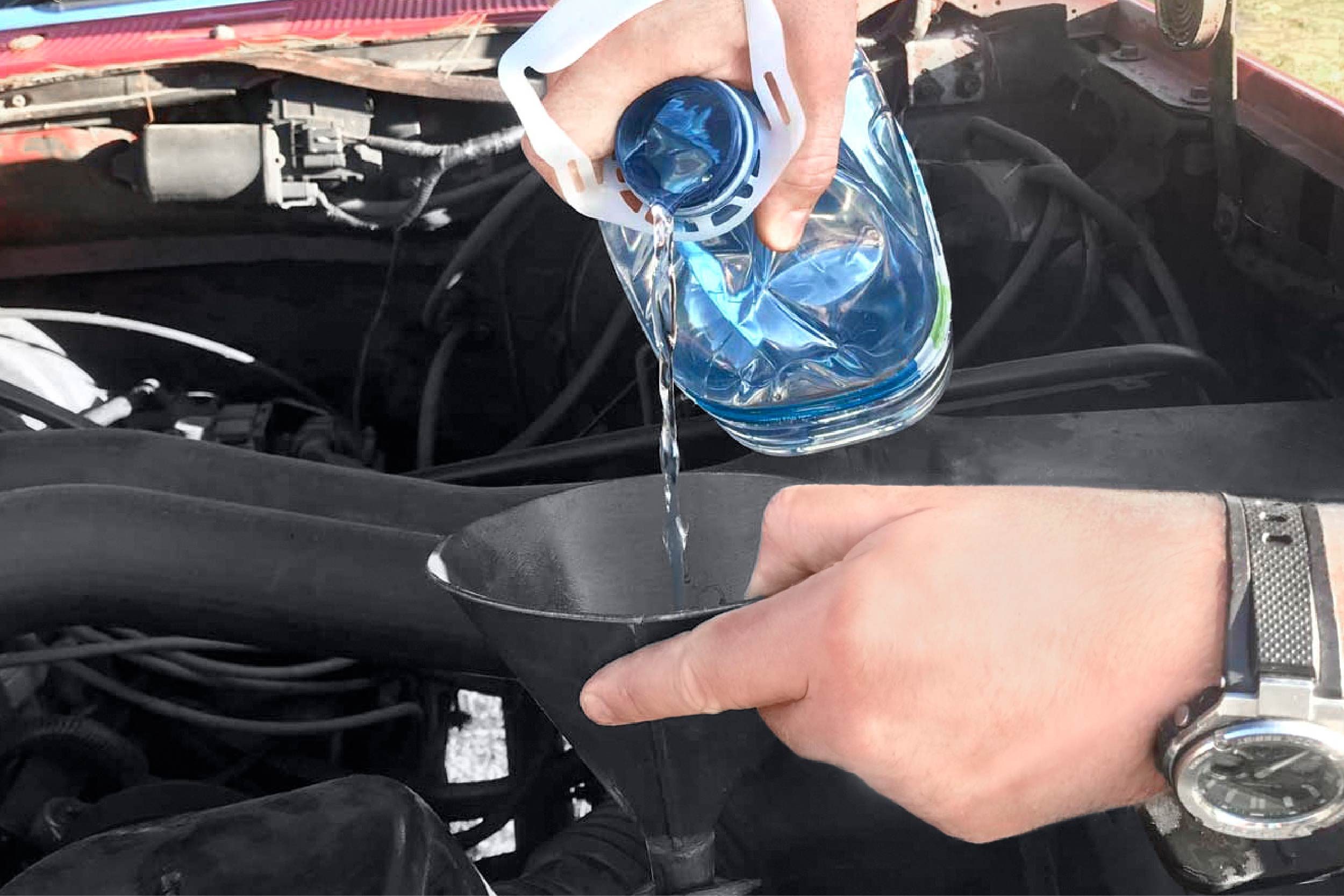 A hand pours water from a clear container into a funnel placed above a car's engine, under a lifted hood in a sunny environment.
