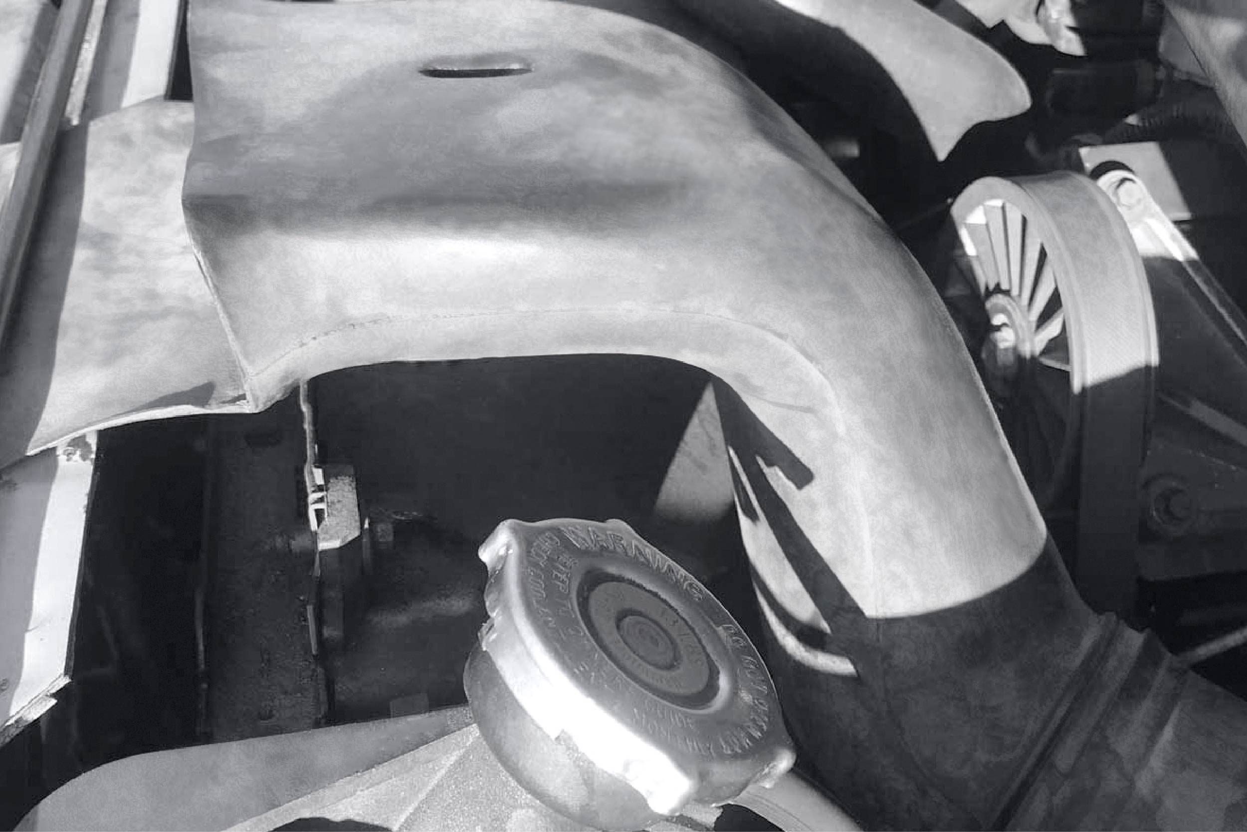 A metal radiator cap sits atop a curved hose in an engine compartment, surrounded by various engine components, illuminated by sunlight.