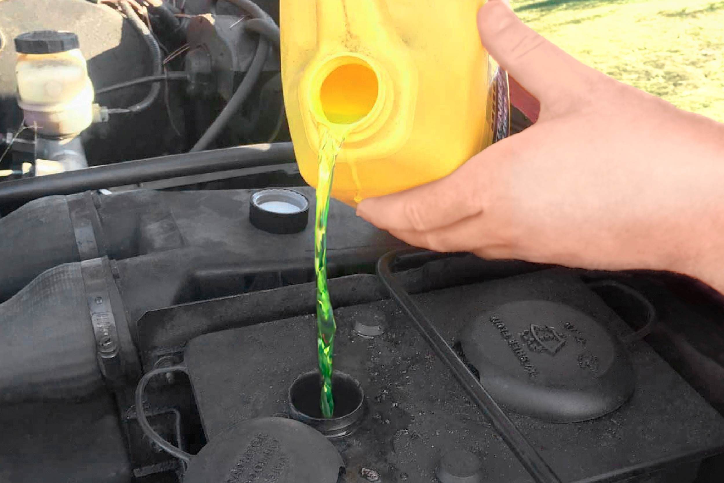 A person pours bright green liquid from a yellow container into an engine's coolant reservoir, surrounded by automotive parts in an outdoor setting.