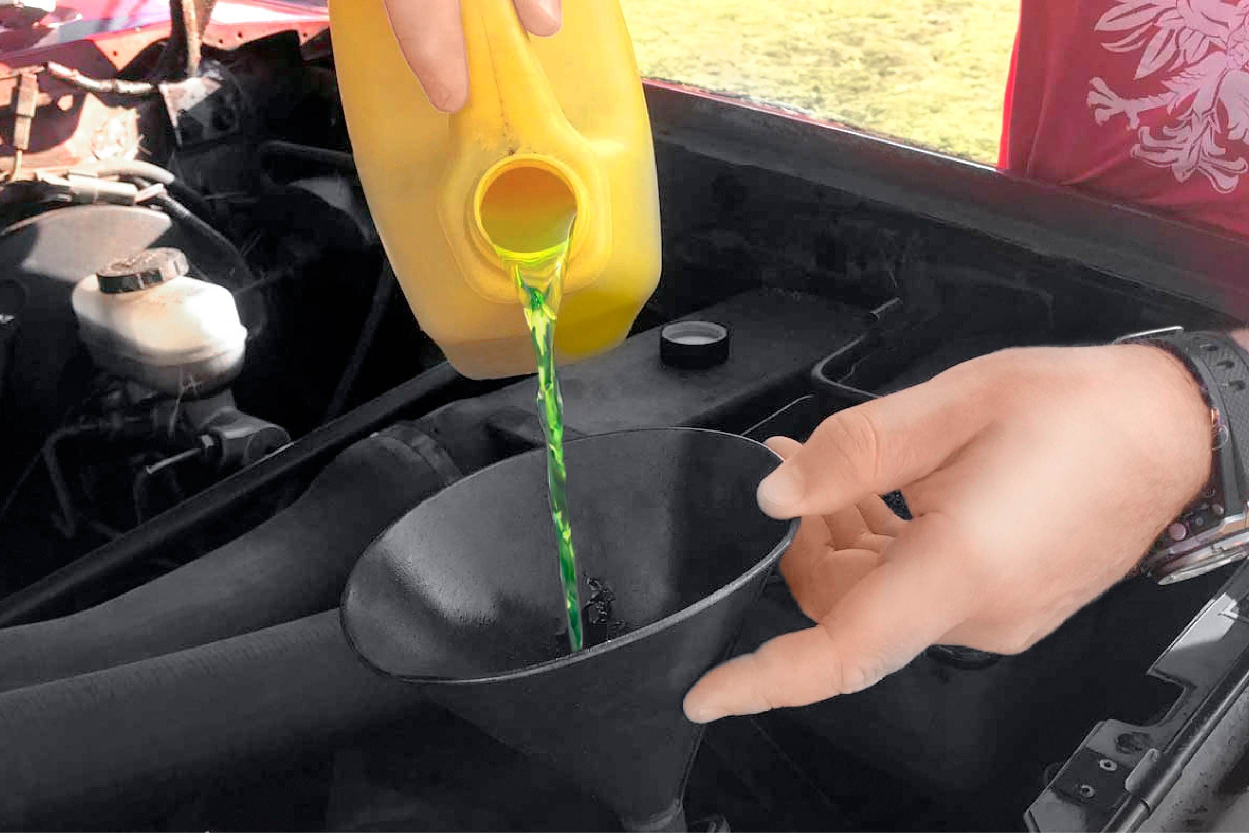 A person pours green liquid from a yellow container into a black funnel situated in an open car engine bay, under natural sunlight.