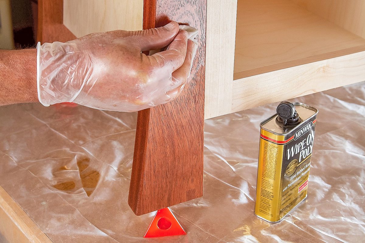 A person wearing a glove applies wood finish to a wooden furniture leg next to a can of wood finishing oil, with the area protected by plastic sheeting;