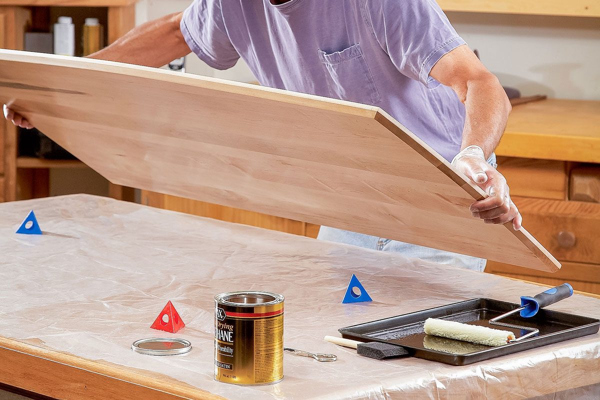 A person wearing glasses and a purple shirt lifts a large wooden board onto a worktable covered with plastic; Wood stain, a paint tray, and small colored wedges are on the table in a wood-paneled room;