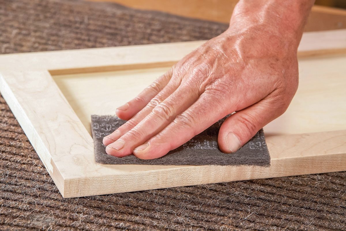 A close-up of a person’s hand sanding the edge of a wooden frame with a sanding sponge, resting on a textured mat;
