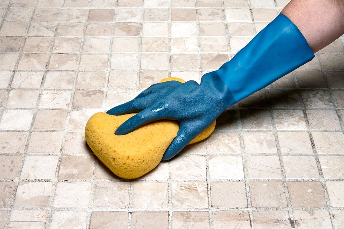 A person wearing a blue rubber glove is scrubbing a tiled surface with a yellow sponge.
