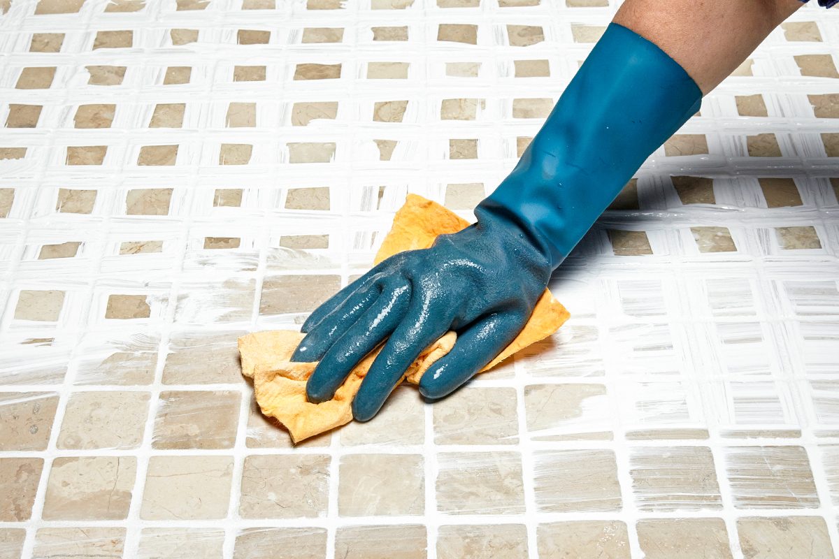 A person wearing a blue rubber glove scrubs tile grout with a yellow sponge on a tiled surface, cleaning the white grout lines.