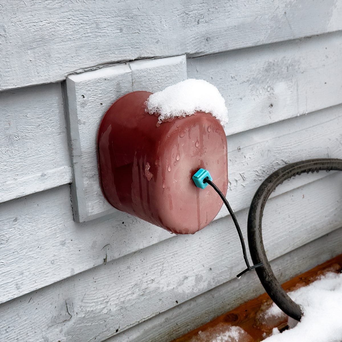 A red outdoor faucet cover with a cable coming out, mounted on a gray house wall. Snow is resting on top of the cover and on the ground below.