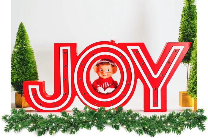 Elf on the shelf in holiday decor. the elf is hidden in the O of a decorative sign that says JOY